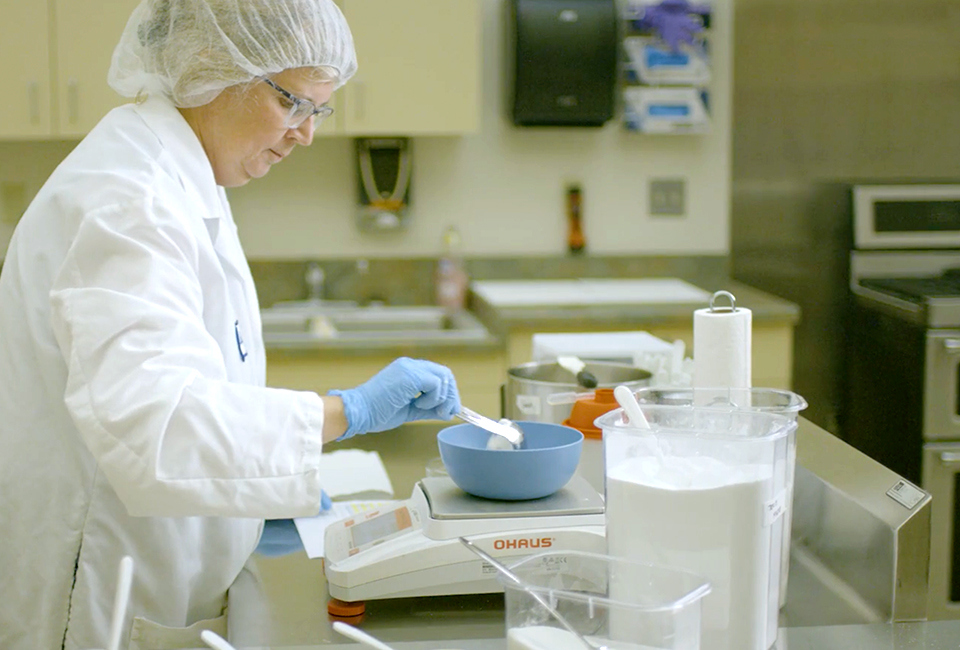 Food scientist in lab coat and gloves weighing scoops of dairy product.